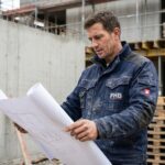 Ouvrier en vêtement de travail FHB sur chantier révisant des plans, veste de travail bleu marine avec poches renforcées